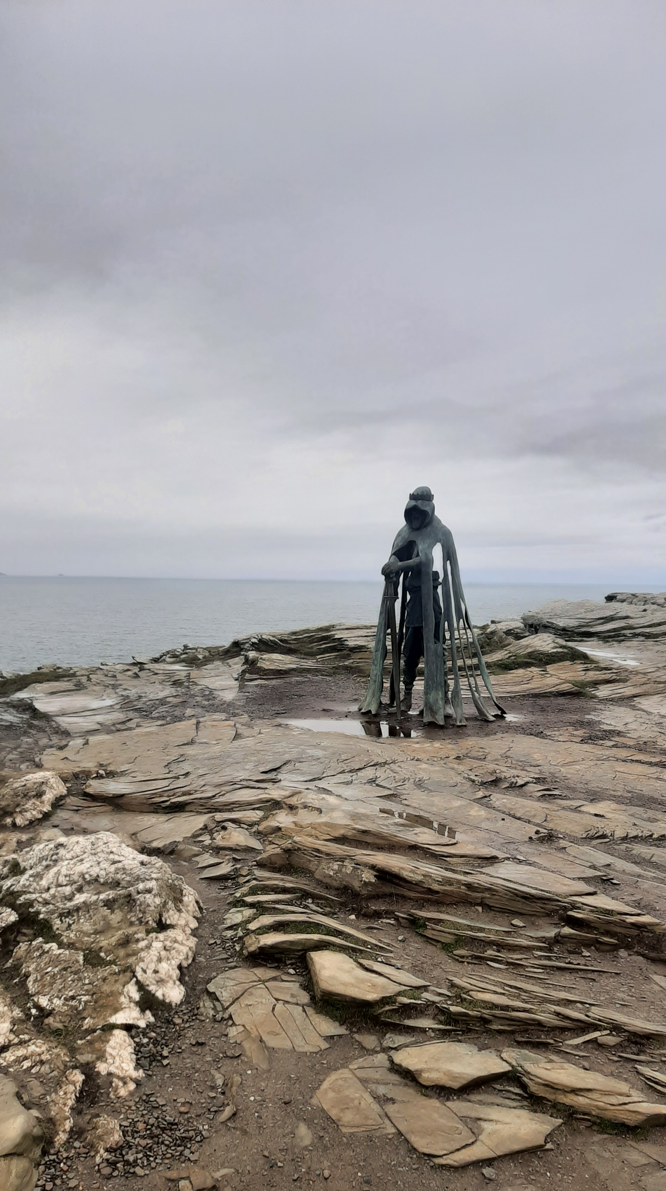 Grey sky, and beige stone clifftop. Atop the cliff is a green statue of a man holding a sword.