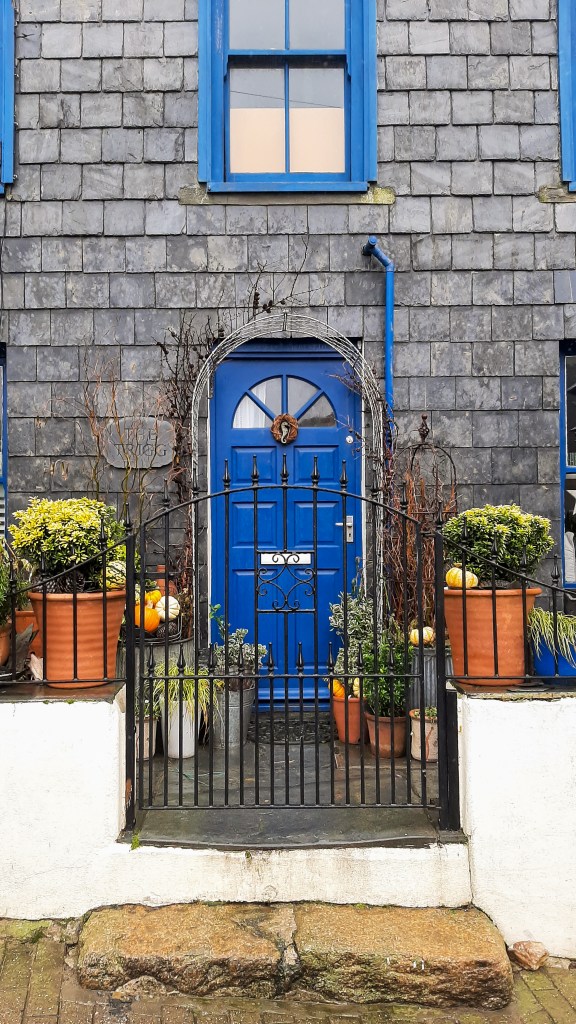 One of the doors of Port Isaac! The doors and windows of this house are bright royal blue, which contrasts against the grey slate. Outside, there are green and yellow garden plants in pots, as well as some small gourds.