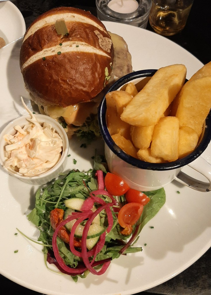 Dinner at The Mote on a white plate. At the top of the plate is a brown brioche bun which holds a burger covered in melted cheese. To the right are thick cut chips in a white pot. At the bottom is a small side salad with cucumber, lettuce, tomatoes, and picked onions To the left is coleslaw in a white tub.