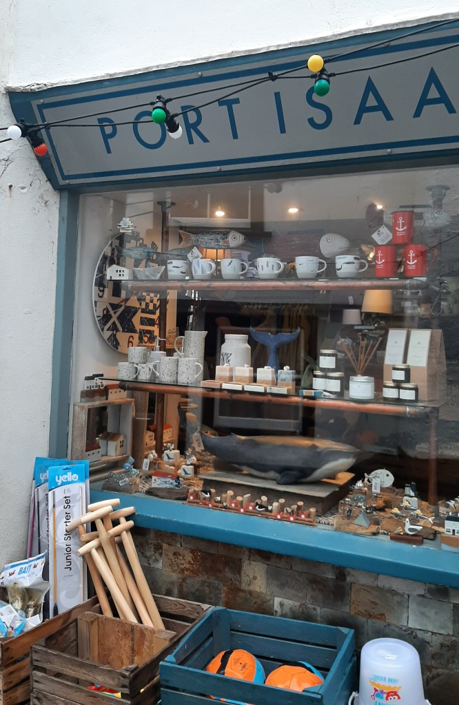 The store front of a Port Isaac shop. The glass window is lined with wooden shelves, and various white, blue, and biege knick-knacks. The outline of the shop front is blue, as the building itself it white with grey brick. In front of the shop, they are selling wooden spades and plastic buckets.