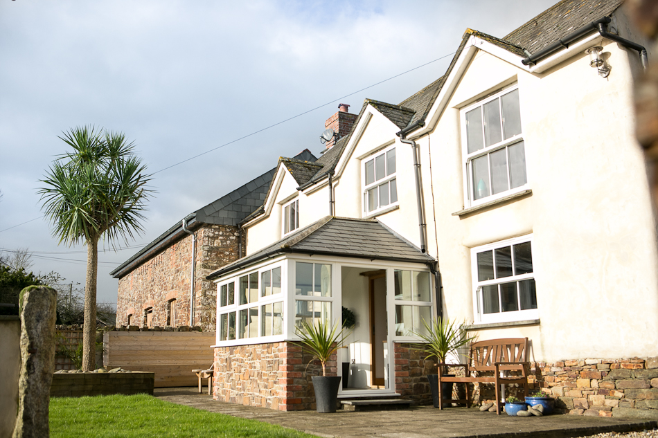 A picture of the Old Farmhouse, Bude. It is a white washed, two story building, with a red brick porch. Infront of the house there is a small patch of green grassm a grey paito, and some all weather plants next to a brown bench.
