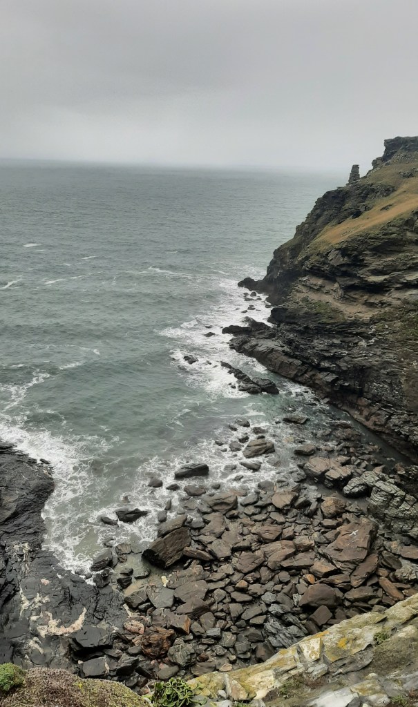 A wild cliffs of Tintagel. The sky is grey and the cliffs are green and grey. The blue-grey sea hits against the rocks that line the inlet.