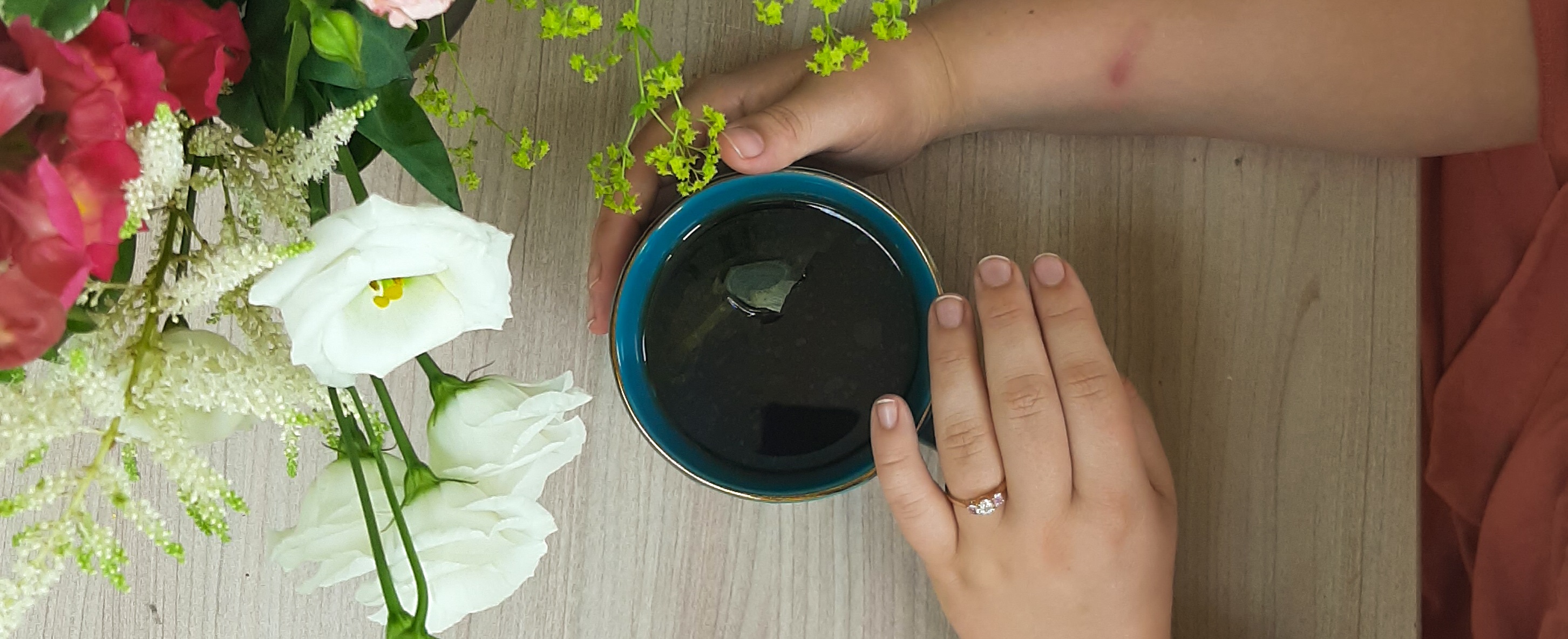 An image of two hands holding a large, blue mug filled the green tea. There are flowers to the left. On the right hand, there is an engagement ring.