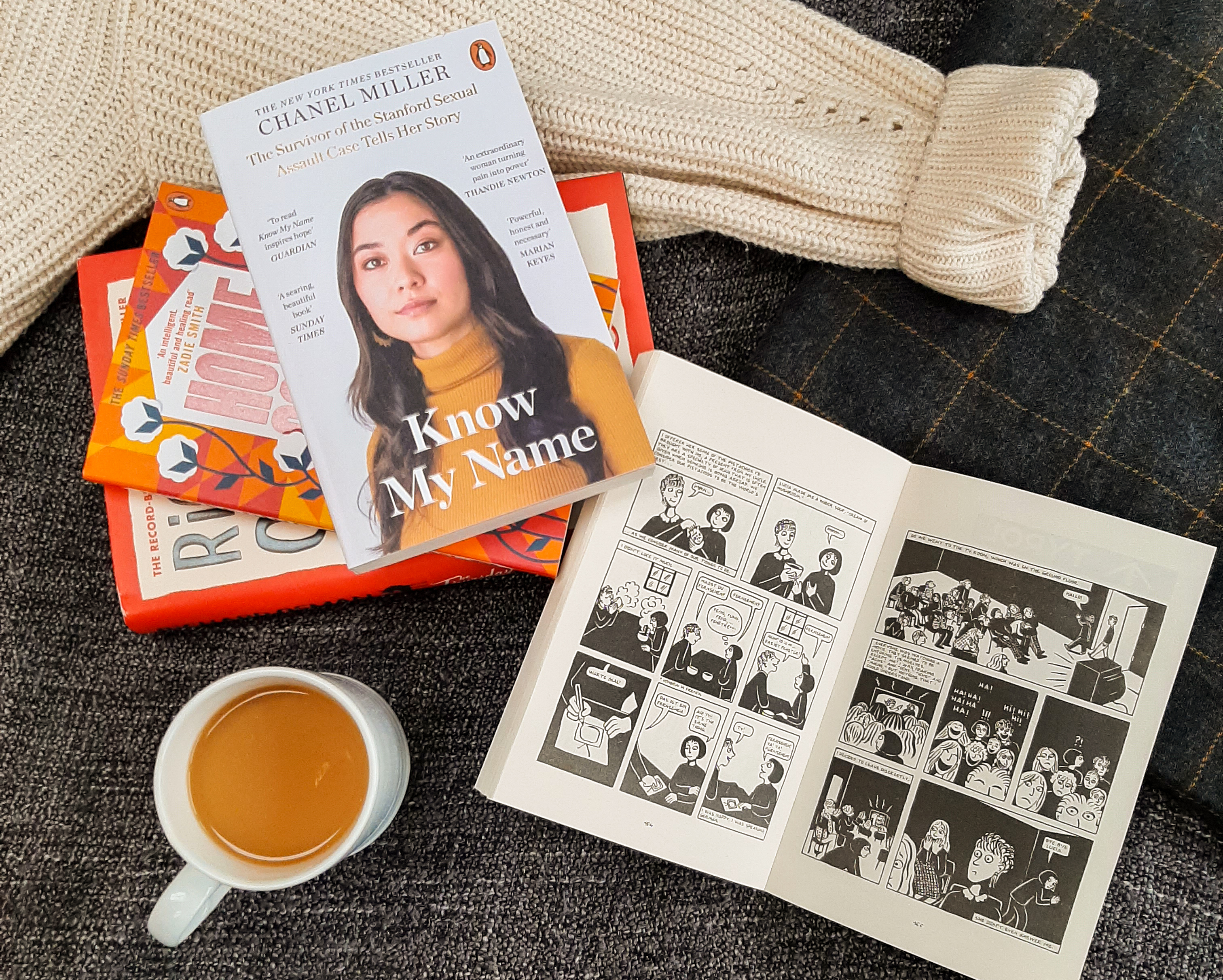 A collection on favourite books of the year. Know My Name by Chanel Miller is at the top of the pile, and Persepolis by Marjane Satrapi is open to the right. The books are lying on a grey blanket with a white jumper. There is a cup of tea next to the books.