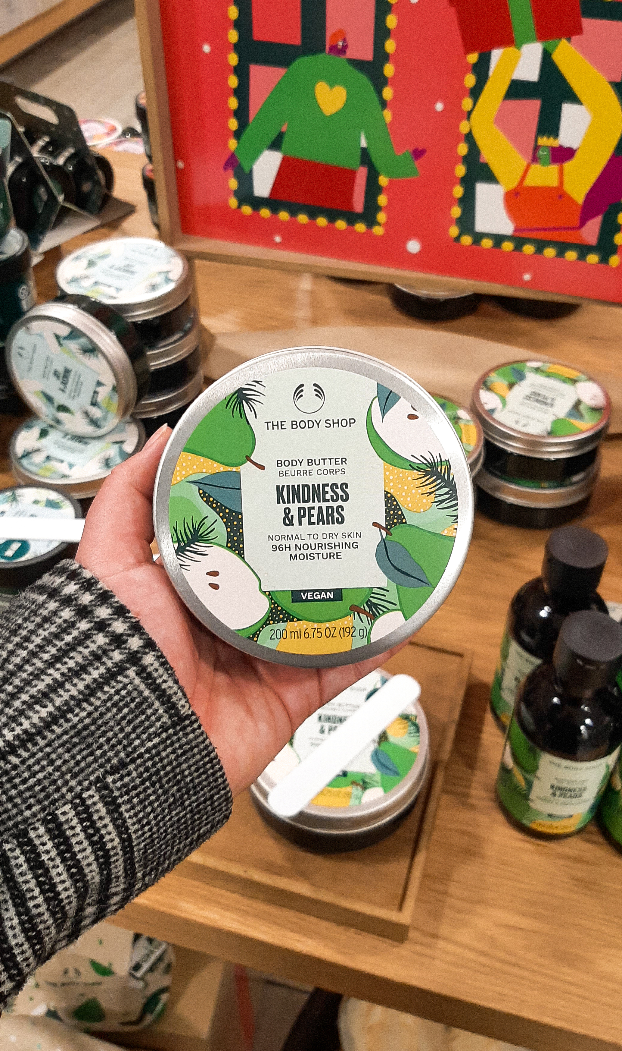 Rosie's hand holding a silver tin of Kindness & Pears Body Butter, with green and yellow print on the tin. The background is of other tins and bottles in The Body Shop.