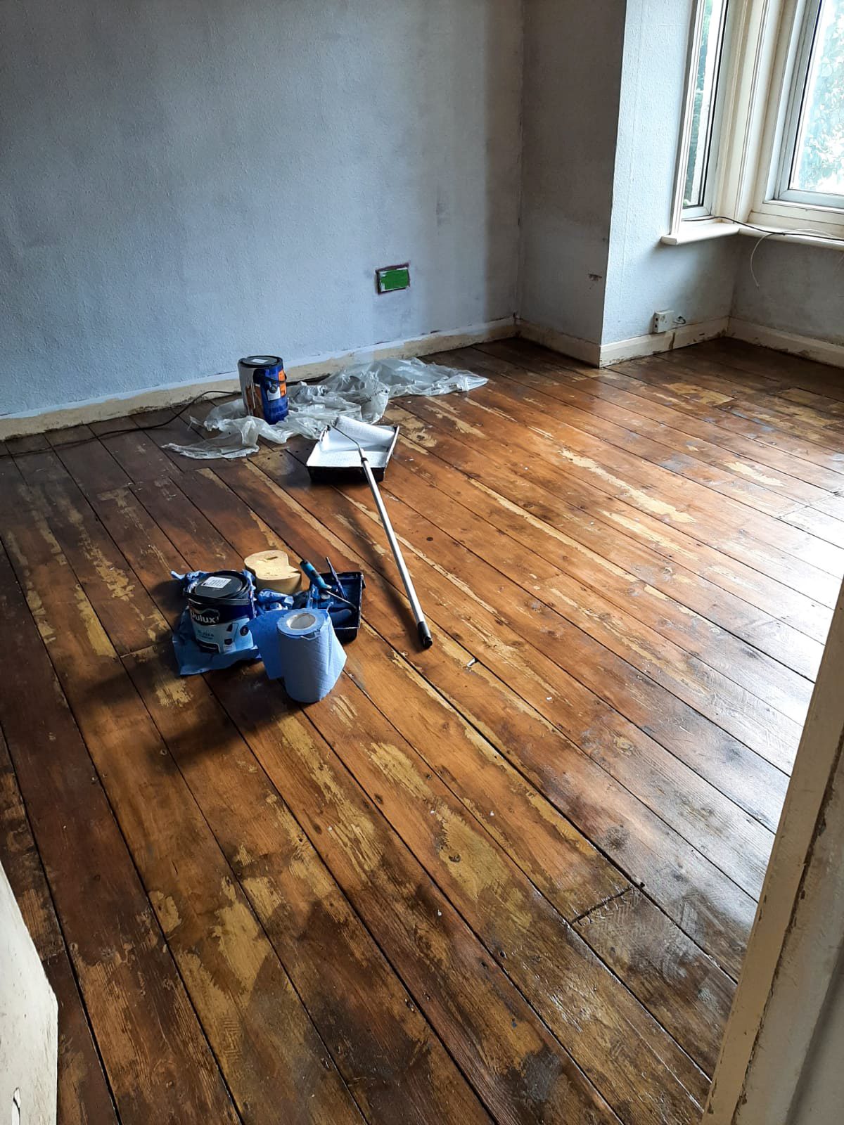 A picture of the living room whilst the floor stain dries. The walls are patchy white, and the floor patchy brown.