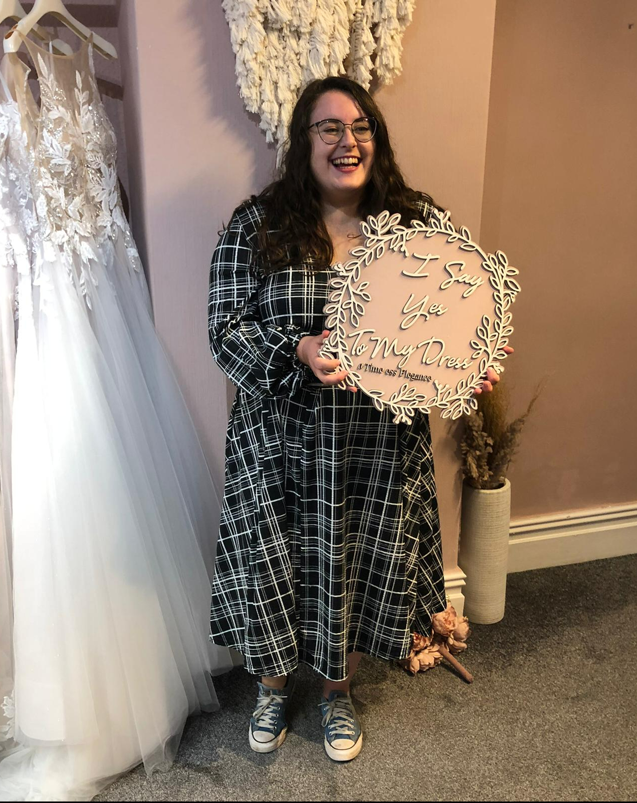An image of Rosie holding a pink circle saying 'I say yes to my dress'. Rosie has dark brown hair, glasses, and is wearing a black and white check dress.