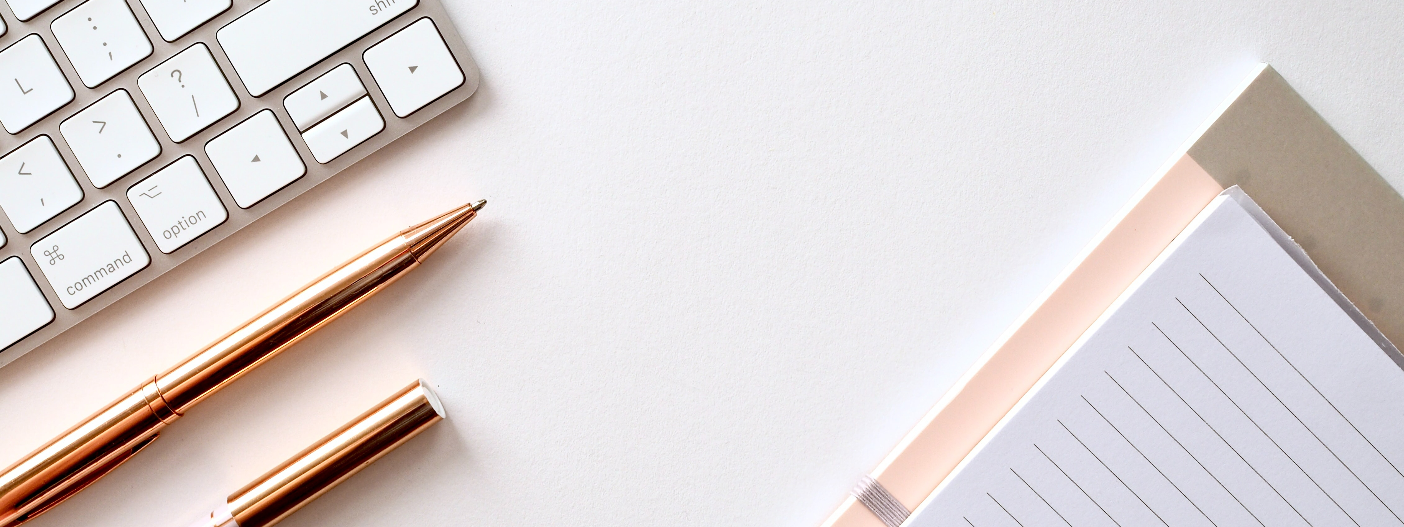 A white background, with two rose gold pens on the foreground. There is also a grey keyboard on the left, and lined paper on the right.