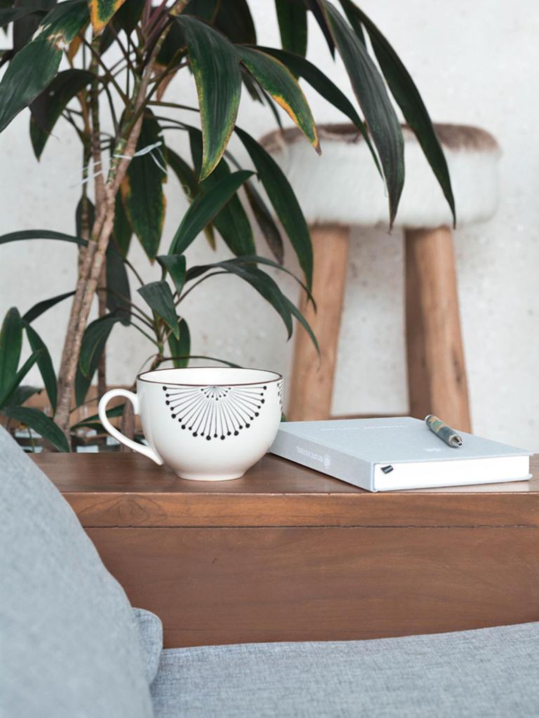 AN image of a white mug and pale blue notebook on a brown wooden sidetable. There is a white and brown stool behind, as well as a green leaf plant.