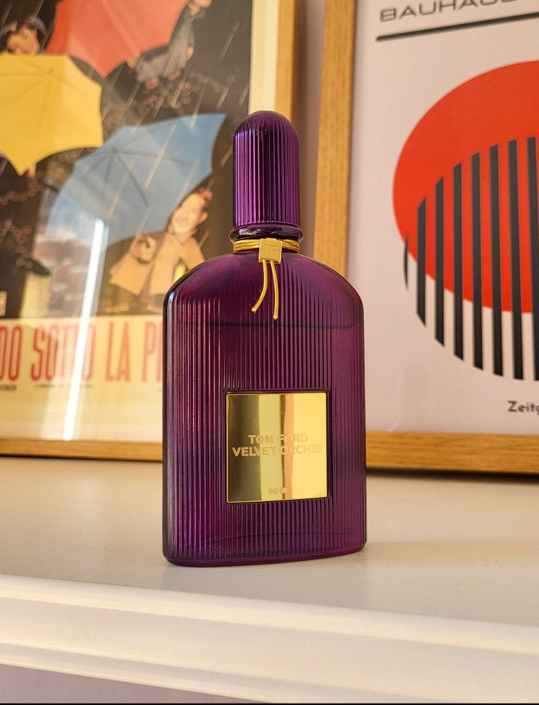 A purple and gold bottle of Velvet Orchid by Tom Ford. It is on a white shelf. Behind it are two colourful prints in light brown frames.