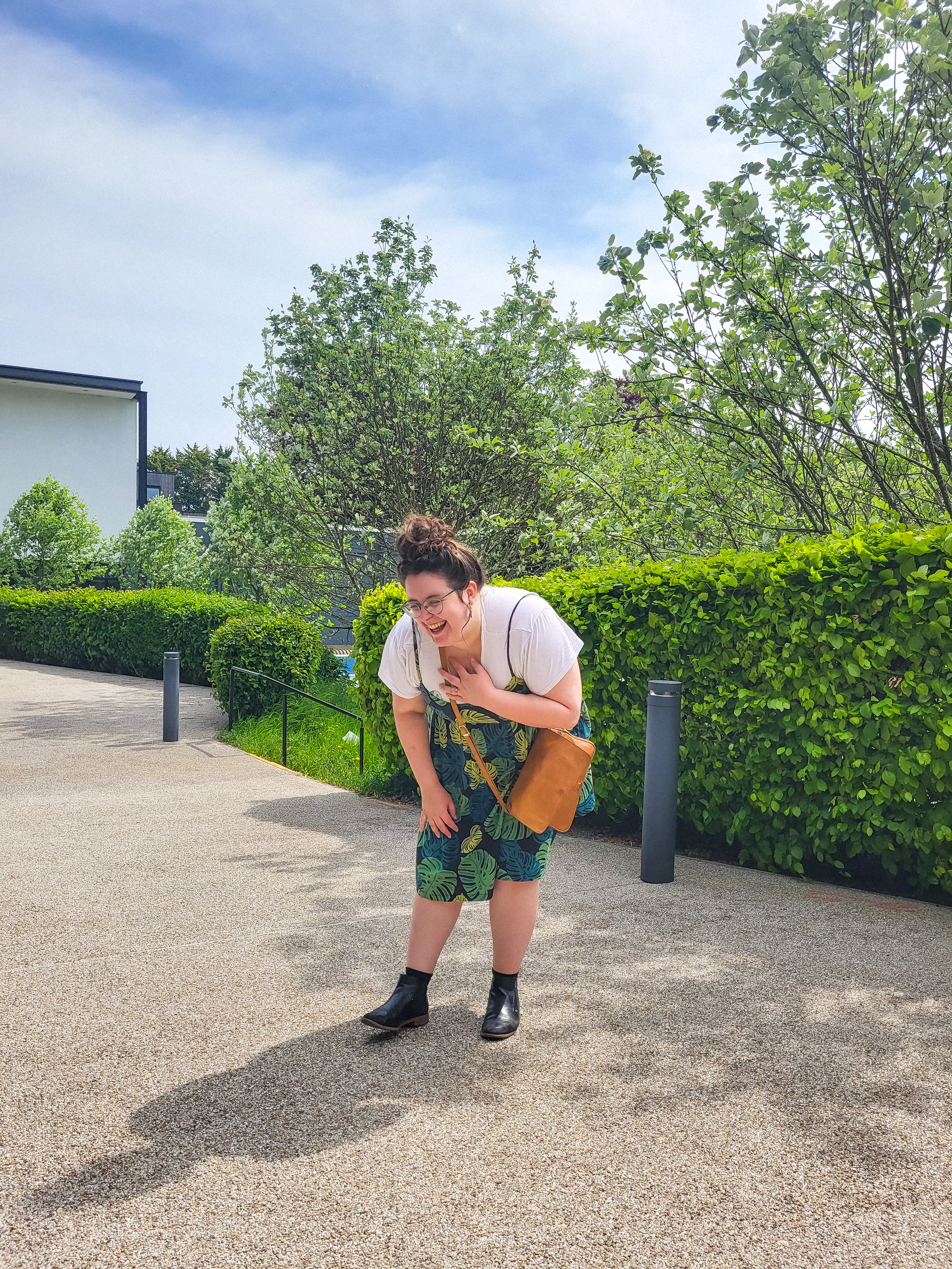 An image of Rosie laughing and clutching her chest. She is a white woman, with brown hair in a bun, and wearing glasses. She is also wearing a white tee, green dress with monstera leaf print, black boots, and a brown bag. The background is green trees and blue skies.