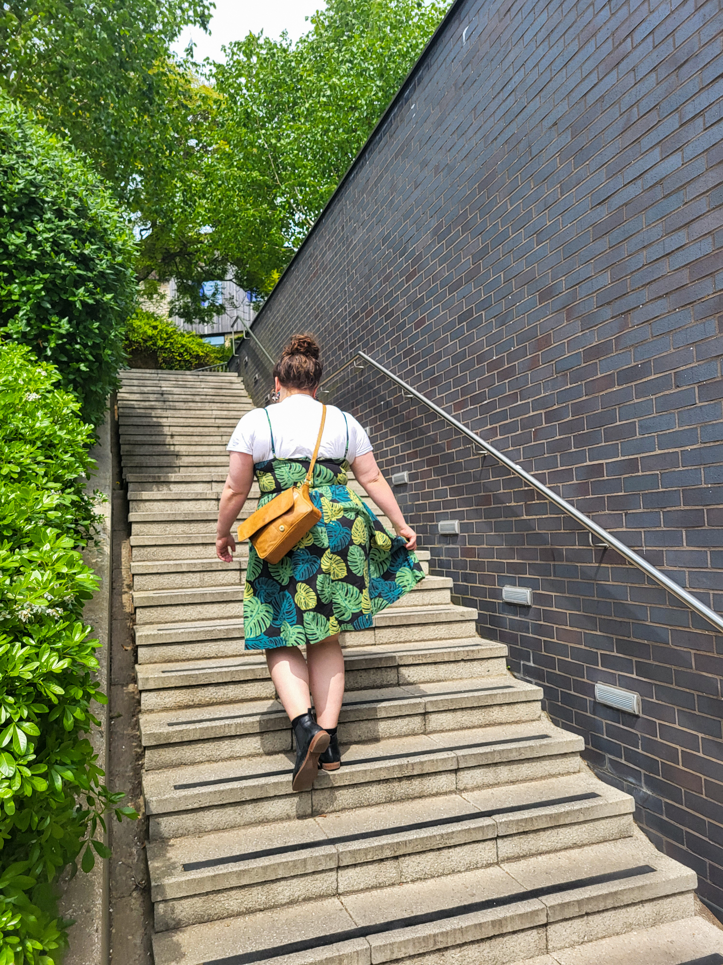 An image of Rosie walking up some concrete stairs. She is a white woman with brown hair which is on her head as a bun. Her back is to the camera. She is wearing a white tee, green dress with monstera print, black boots, and a tan satchel. There is a black brick wall to the right and green bushes to the left.