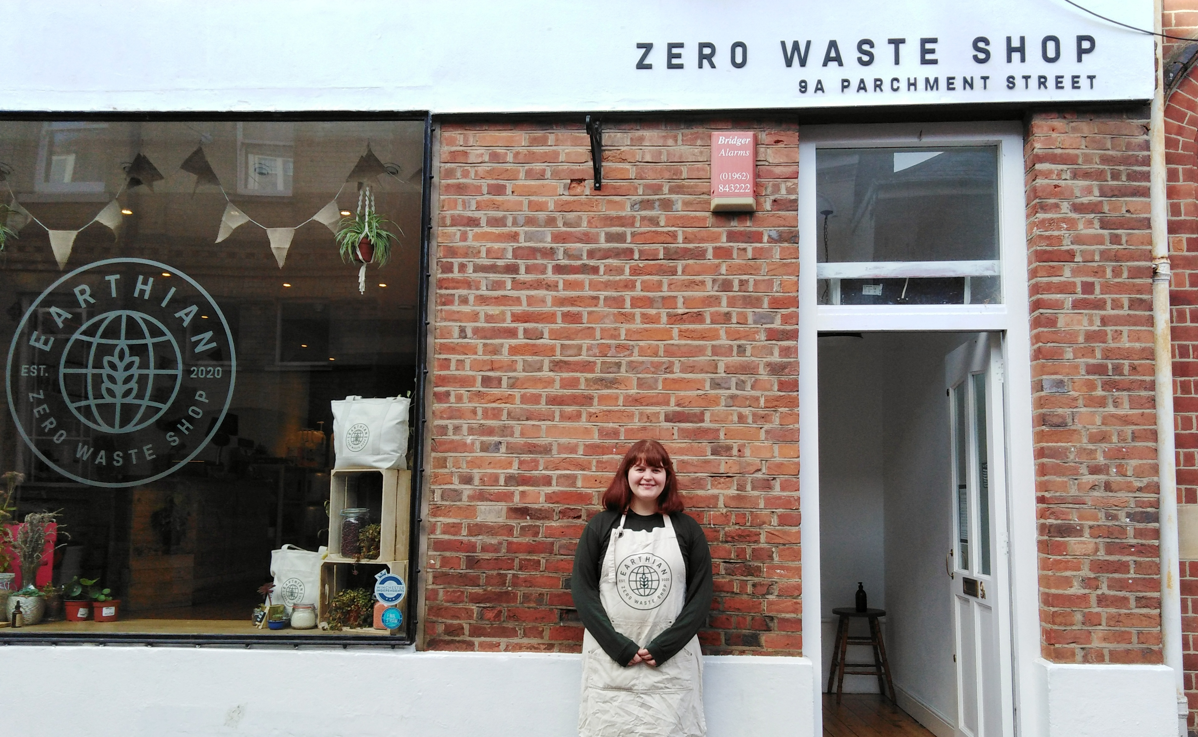 A red brick wall and a glass window saying 'Earthian' A redheaded Caucasian woman, Katie, stands in front of the red brick.
