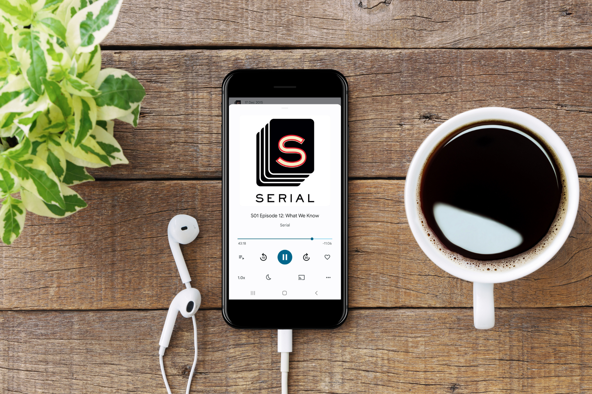 An image of a phone on a wooden table, with the Serial podcast. There are plants on the left, and a black coffee on the right. There's also white headphones on the table.