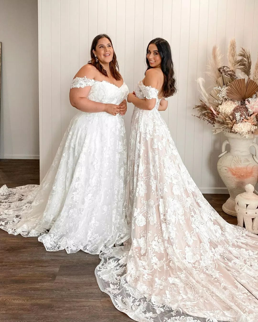 An image of two woman in the same floral, white wedding dress. The one on the left is curvy, the one on the right is thin. The are both brunettes.