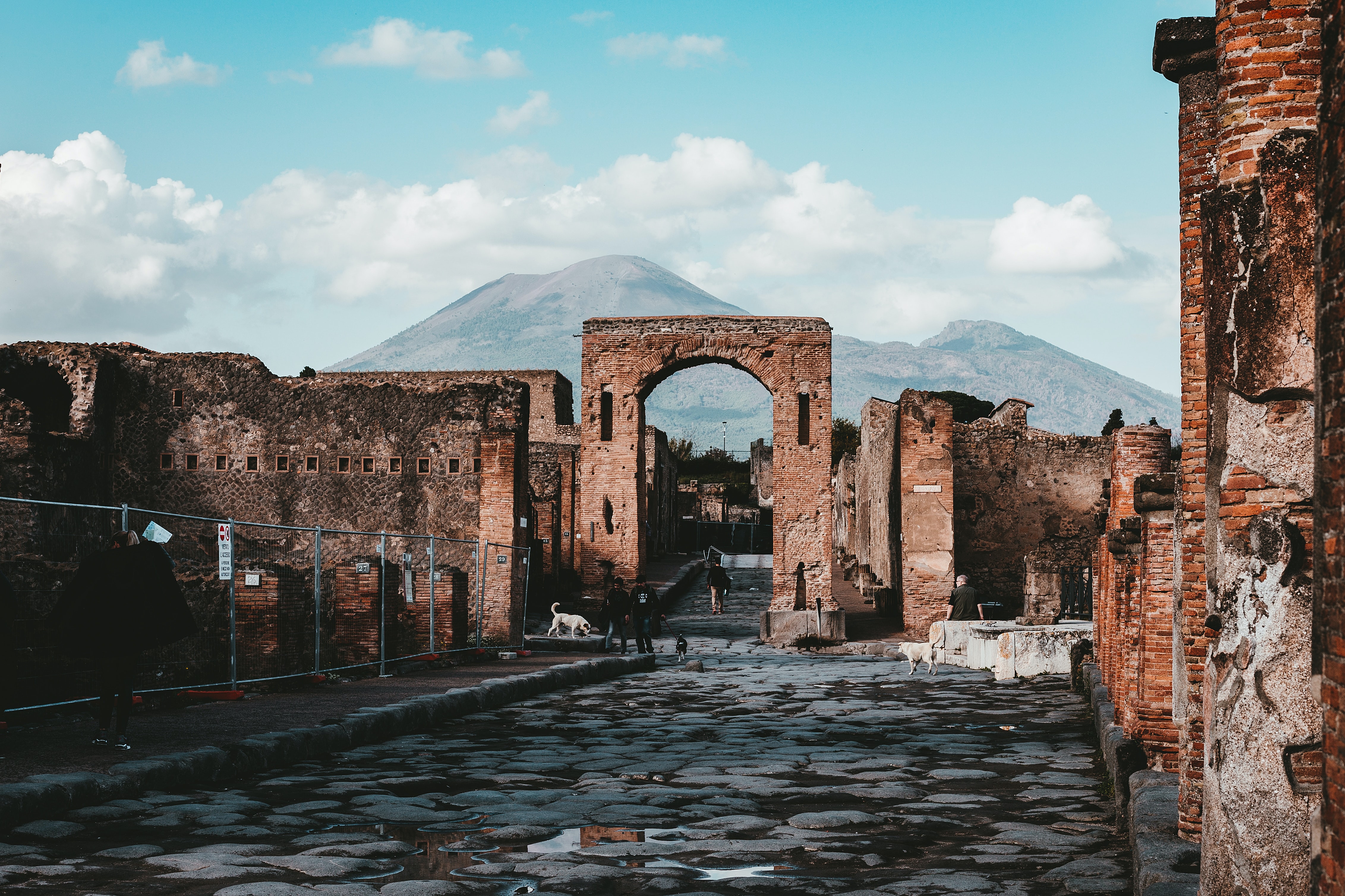 The brown ruins of Pompeii with the green and brown hills of Vesuvius in the background. The sky is blue and cloudy, and the photo centres on a brown arch