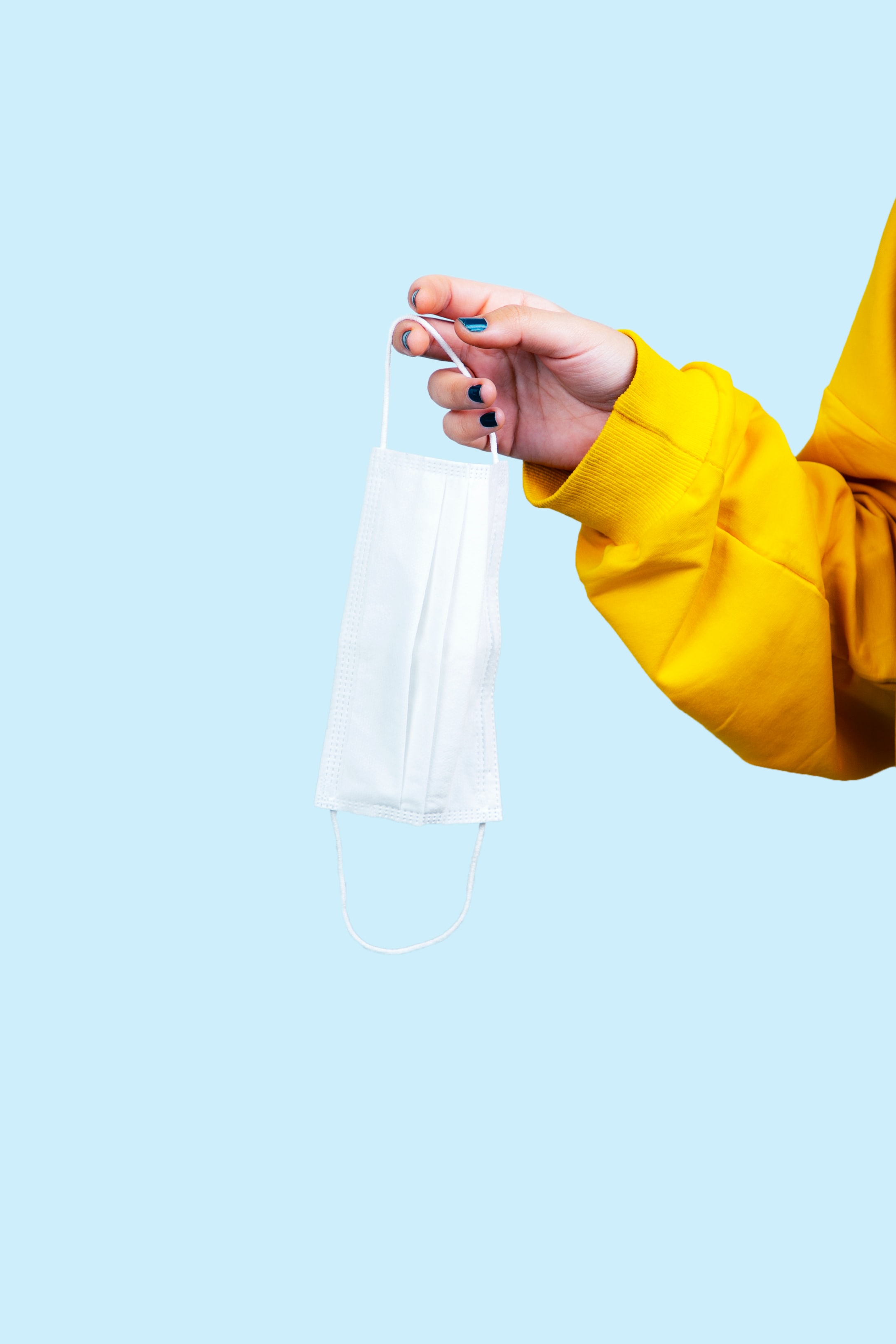 A light blue back drop, where a white arm in a yellow sweatshirt is holding a white COVID mask.