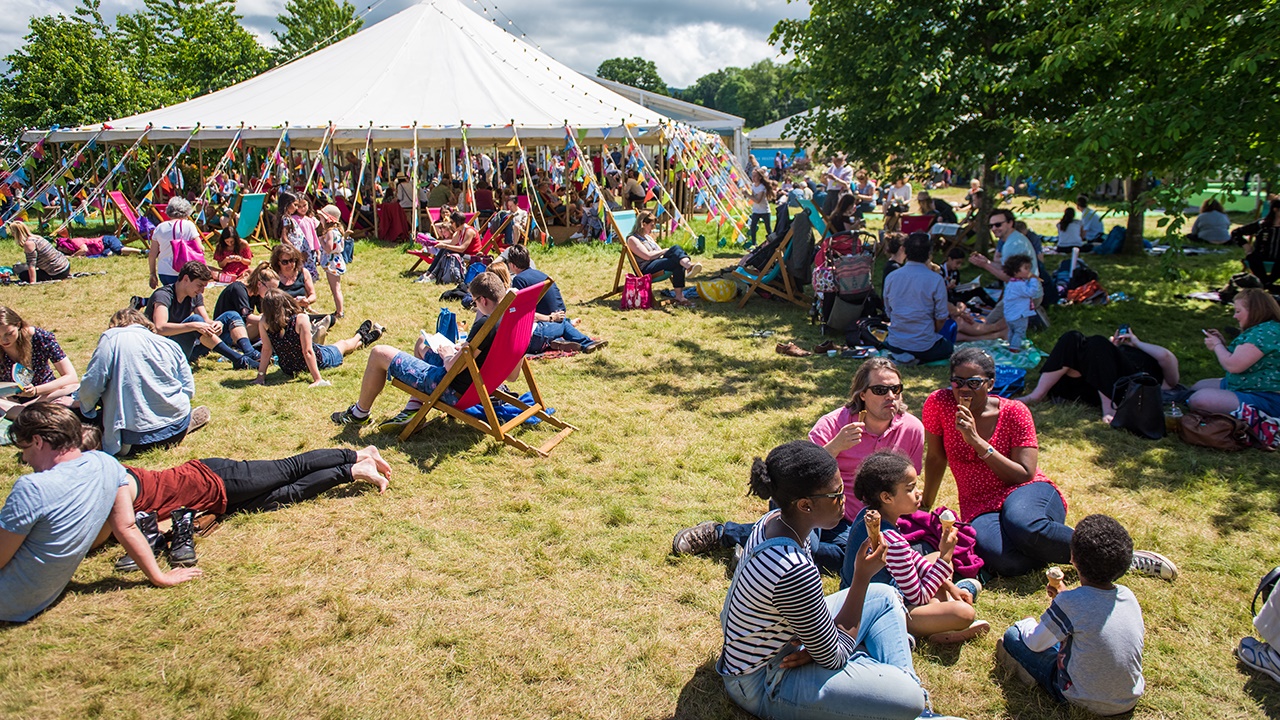 People sitting on the yellow green grass or on chairs at Hay Festival. In the background, there is a white tent with colourful flags.