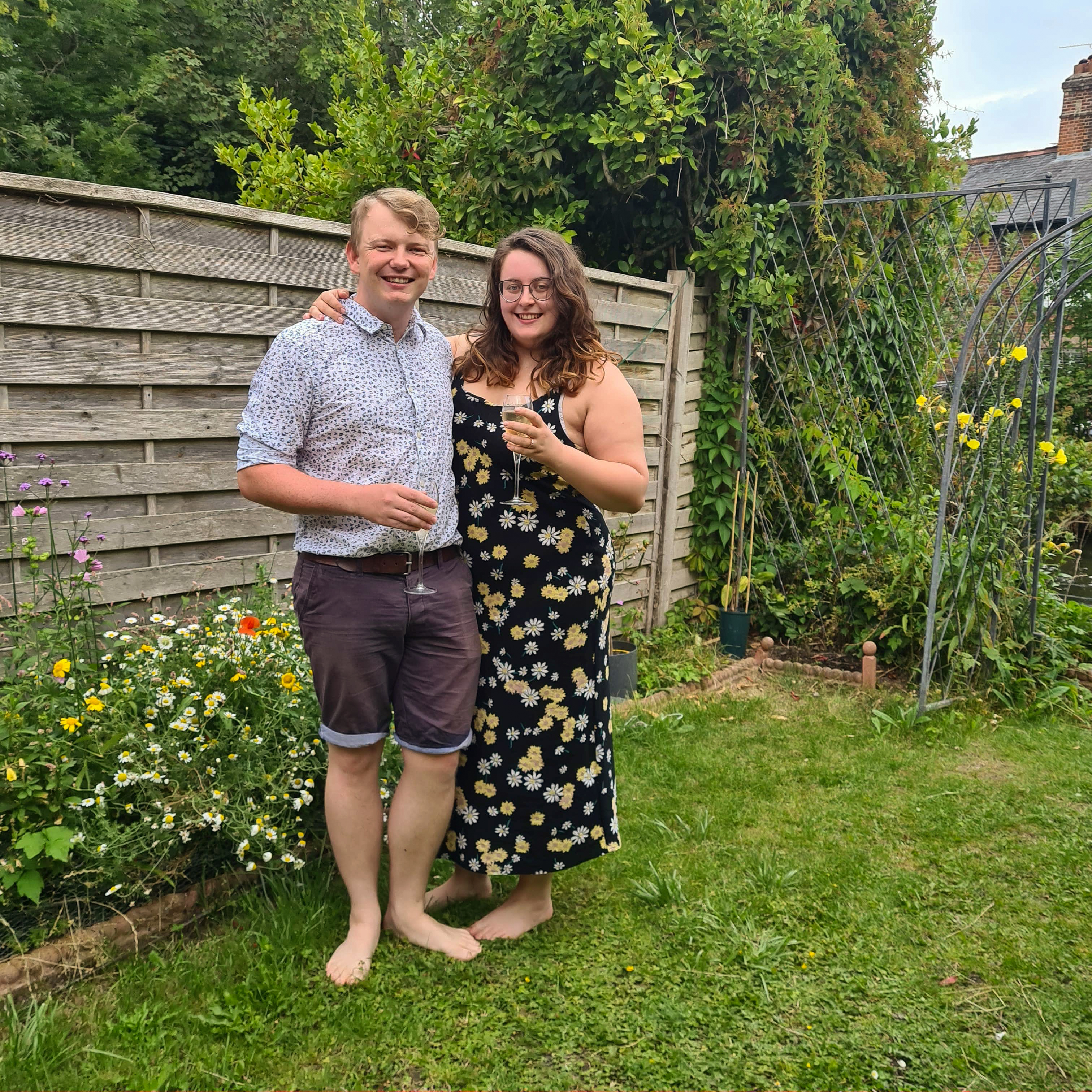 An image of rory and Rosie together. They are both white and are of similar height. Rory is on the left and is wearing a light blue shirt with dark shorts. Rosie has brown hair and is wearing a maxi black dress with daisies. They are outside, standing on green grass, and holding glasses of champagne.