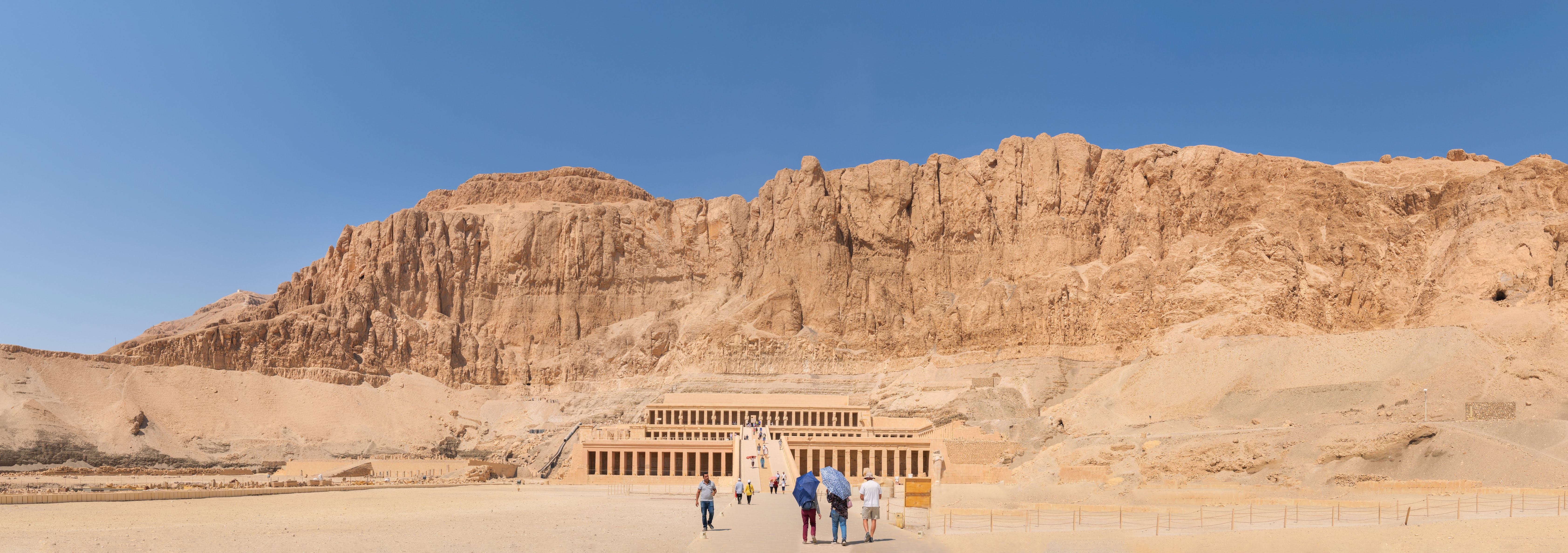 An brown-orange cliff, over looking a stone temple built into it. The sky is blue and the floor is sand. A group of people walk up to the temple.