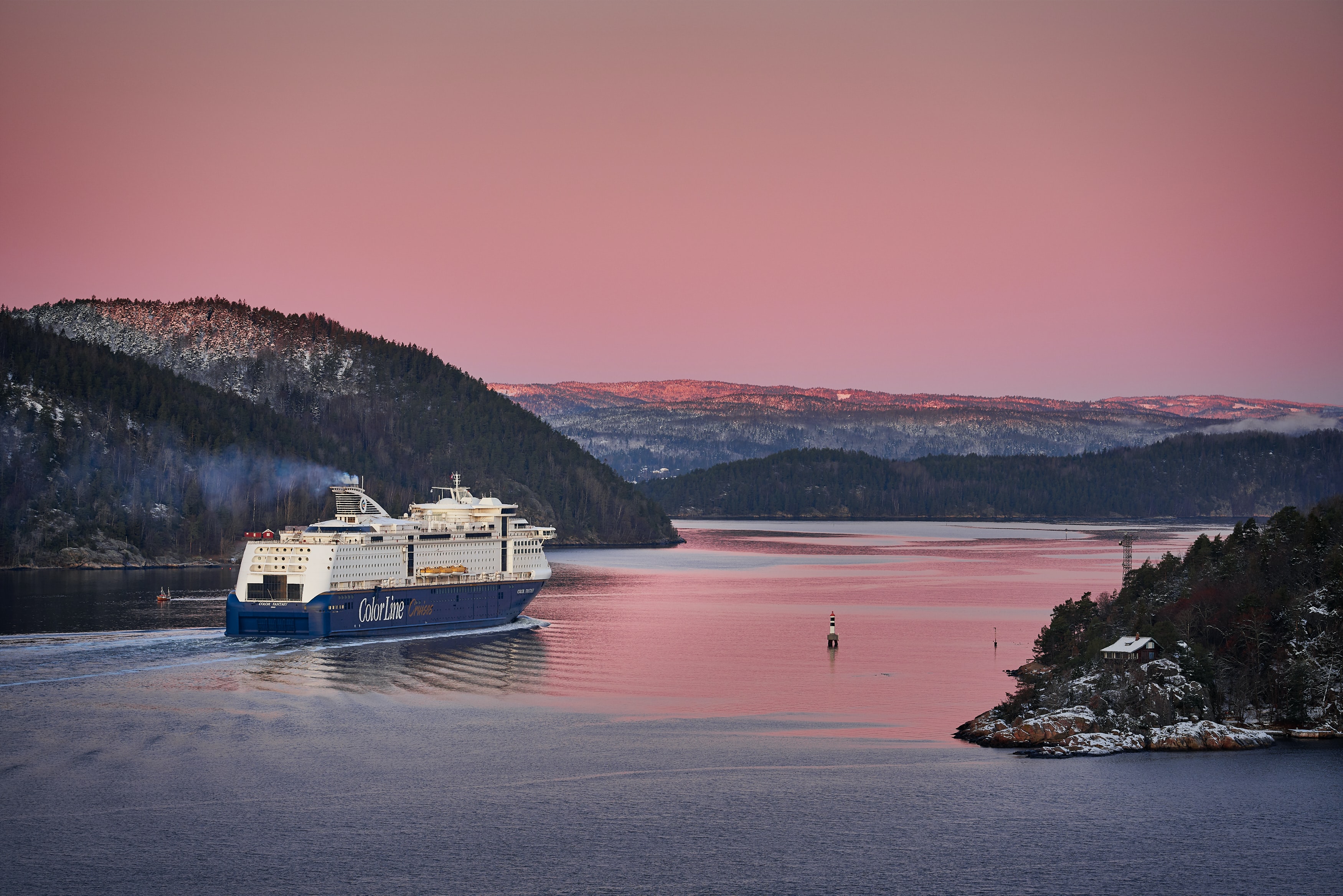 A pink sky, grey snow capped hills, and dark blue river. There is a white and blue cruise boat on the river.