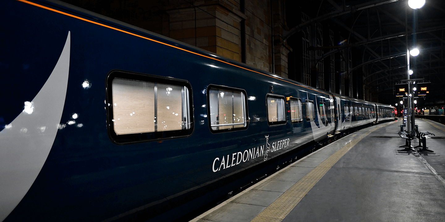 An image of a dark blue train carriage that says 'Caledonian Sleeper' in all white font. It is night time and is at a grey platform