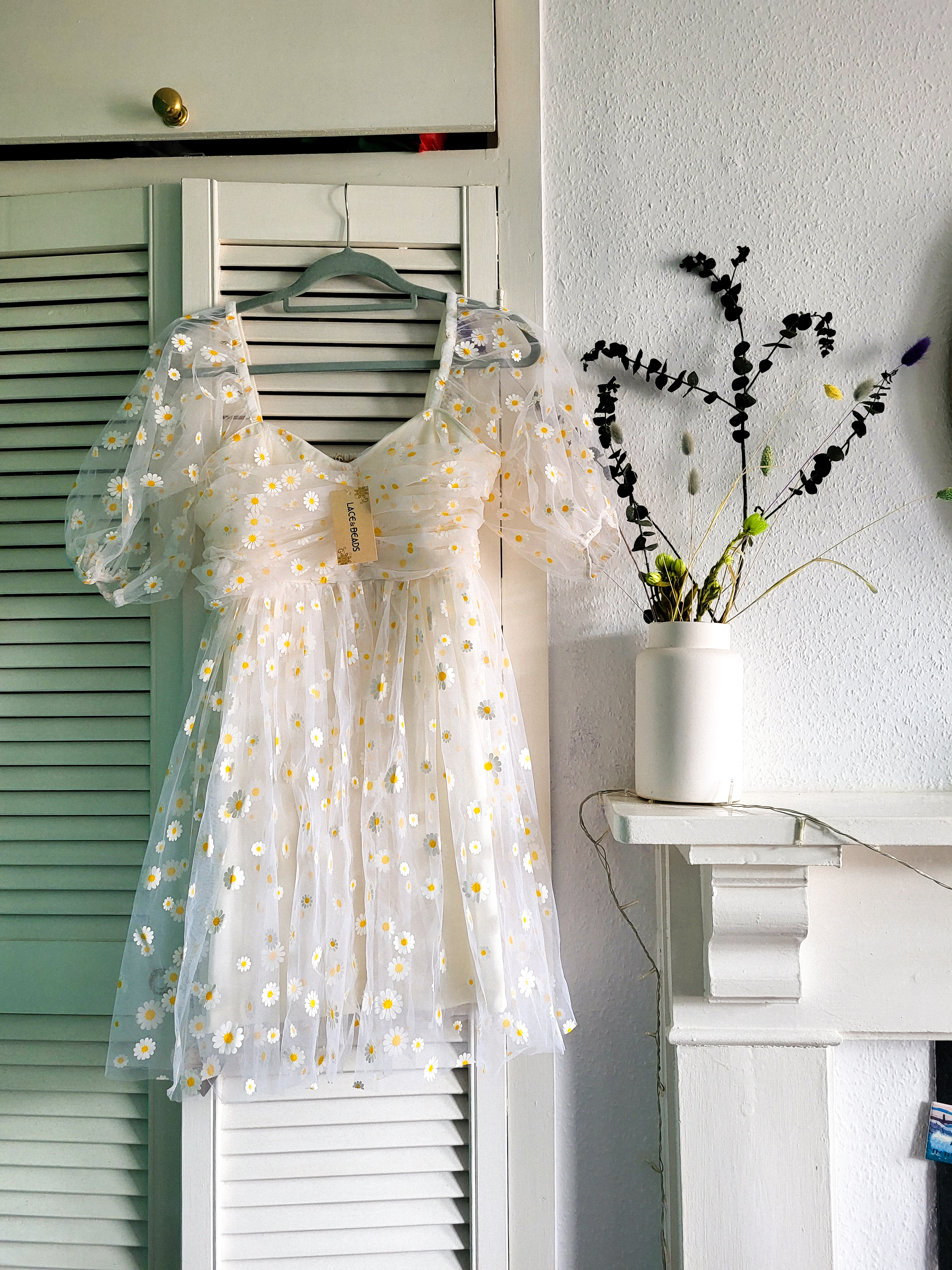 An image of a white dress hanging on the door of a white cupboard. The dress is made of tulle and is printed with daisies.