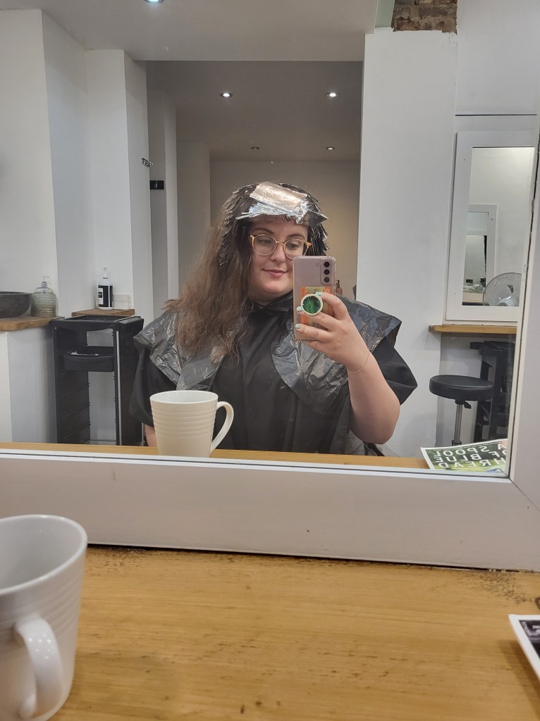 Rosie sat in a hair salon. She has brown, brushed out, frizzy hair that has silver foils in. She has glasses, and is holding her lilac phone up to the mirror to that the photo.