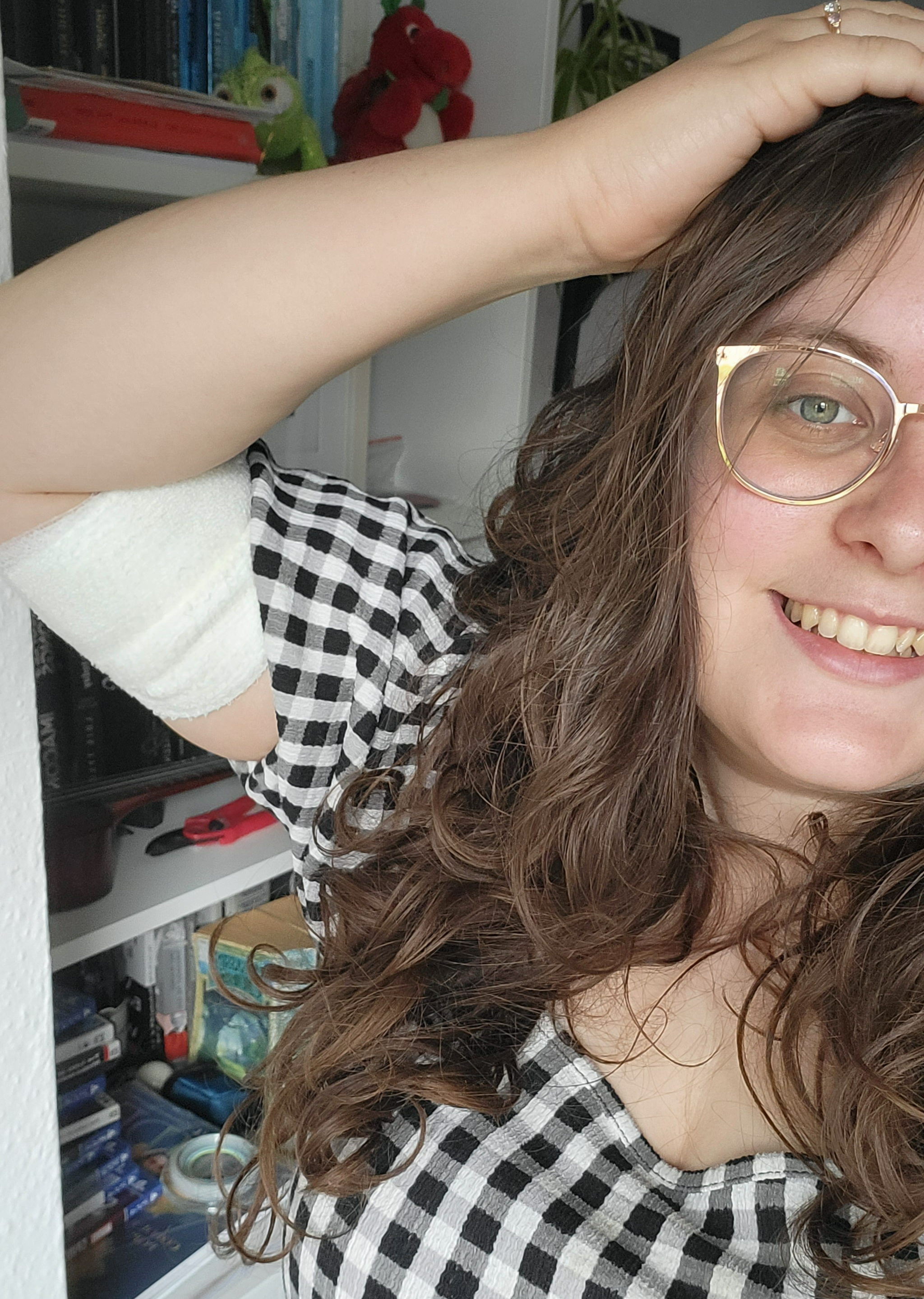 An image of Rosie looking to the camera, with a white bandage on her upper arm. Rosie is a white woman with long curly brown hair and gold rimmed glasses. She is wearing a white and black check dress with a square neck and puffed sleeves. Half of her face is hidden.