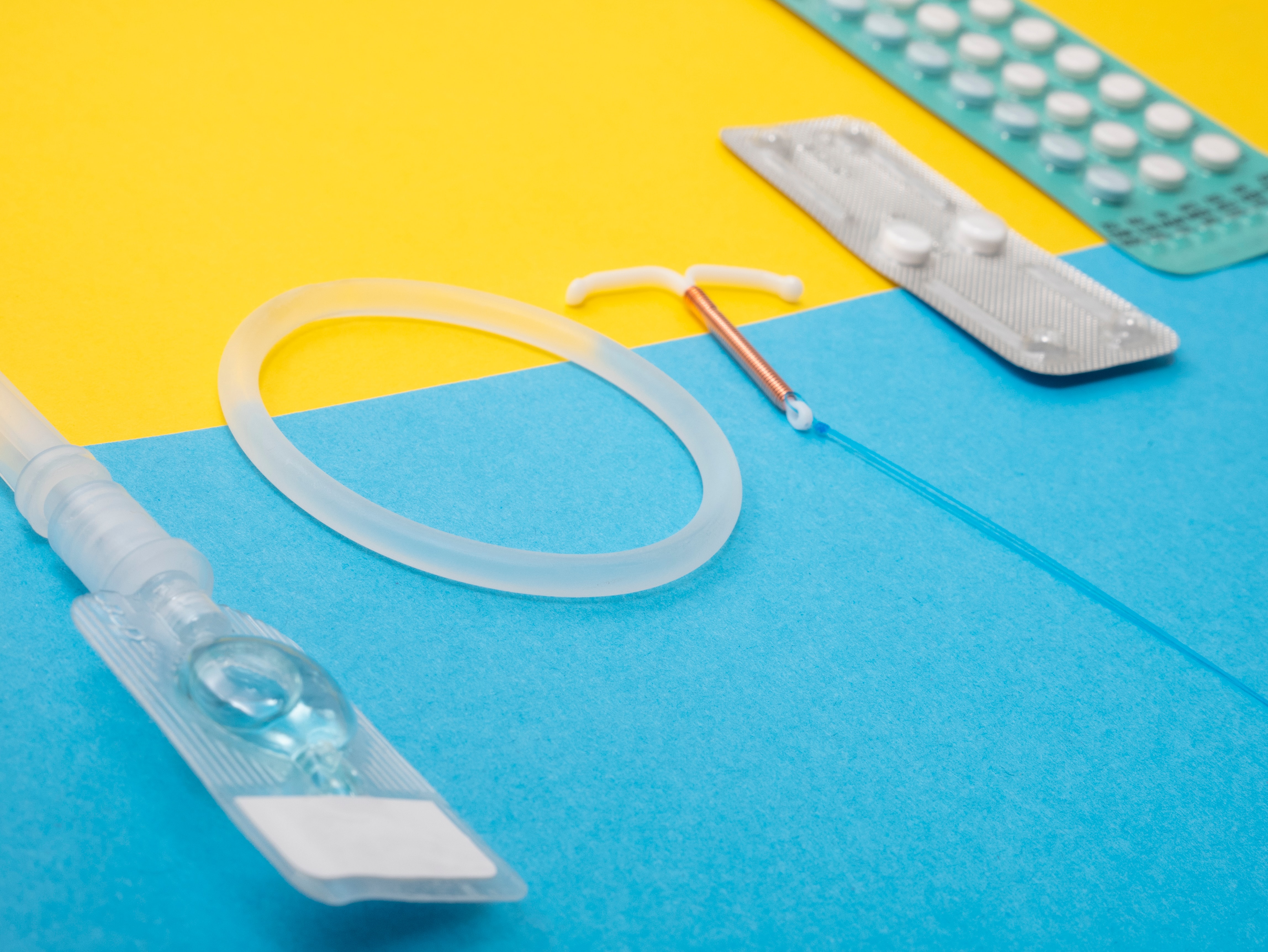 An array of contraceptives on a yellow and blue background. Left to right - needle and fluid; clear ring; copper coil; two white pills in a silver sleeve; white pills in a blue sleeve