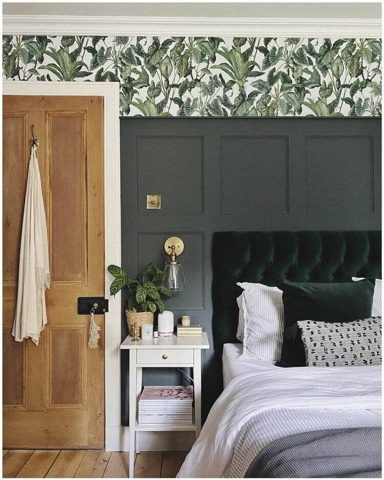 An image of a bedroom. On the left is a light wood door, on the right is a green velvet bed. Between them is a white bedside table with knick-knacks. Behind that, is a green panelled wall, with tropical leaf wallpaper above.
