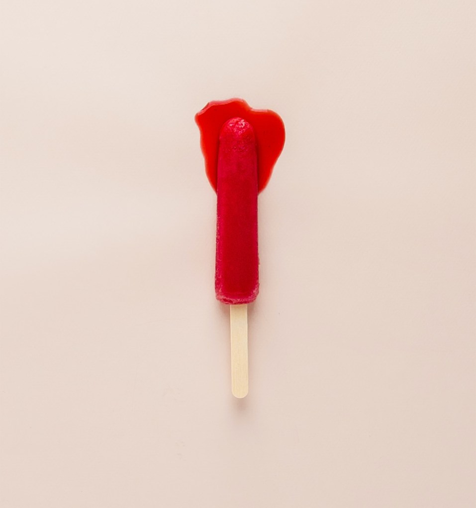 A red lollypop melting on a pale pink background. It evokes the image of a bloody tampon.