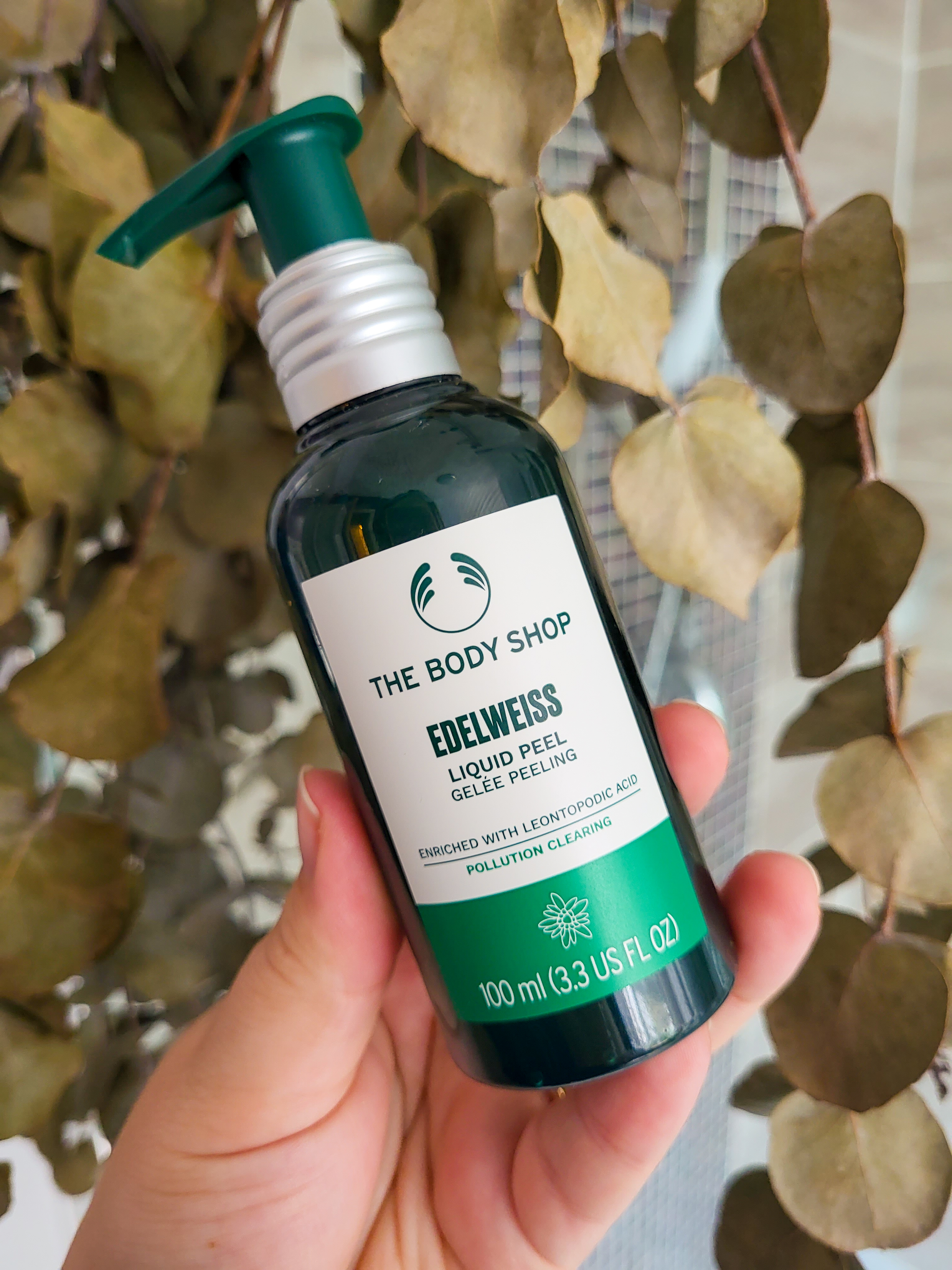 Rosie's hand holding up the green and white pump bottle of the Liquid Peel. The Body Shop logo is on the top of the bottle, a pump is above it. The background is dried eucalyptus.