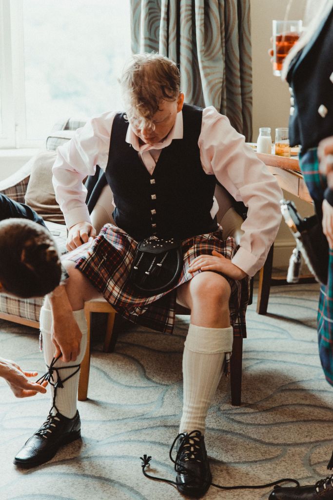 An image of Rory sitting on a chair, legs a bit apart, hands on knees. He is a white man with light brown hair, and is wearing a black waistcoat, light pink shirt, red tartan kilts, white knee socks, and black shoes. Hands to the left of the image are tying his laces. 