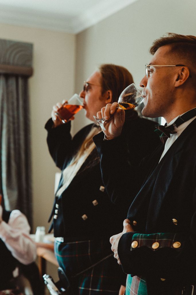 In focus, on the right of the image - Joey, a young white man with glasses, is sipping from a wine glass with whiskey init. Joey is wearing a tartan bowtie, a black jacket with gold buttons, and a tartan kilt. More out of focus, and to the left of the image, Alex is doing the exact same thing, in a similar outfit. Alex is white, wearing sunglasses, and has long brown-red hair. They are drinking a pint. 