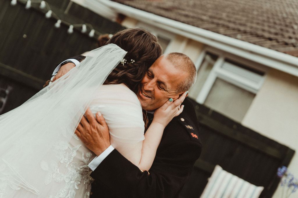 Rosie, brown hair, hugs her father on her wedding day. She has tulle sleeves, and he has a black jacket on. You can see his face and he is smiling. 