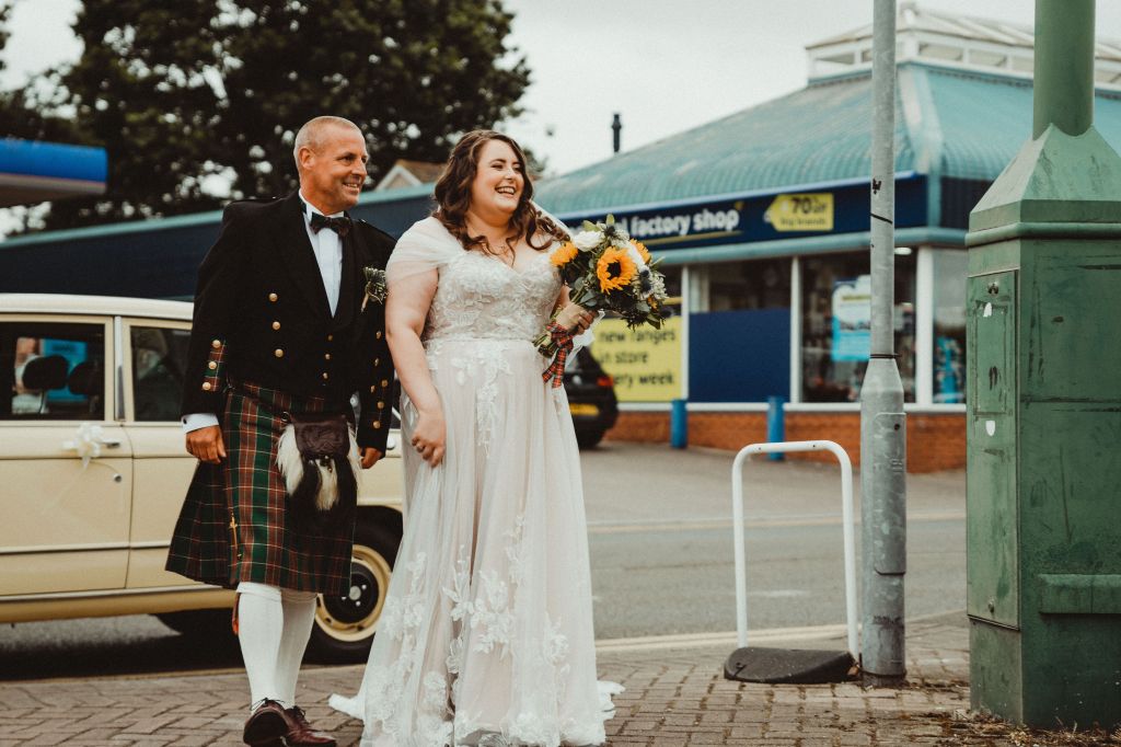 Rosie and her dad walking. He is to the left of the image, and is wearing a red and green traditional Welsh cilt. Rosie is to the right, with wavy brown hair, a white and lace wedding dress, and is holding a bouquet of flowers. 