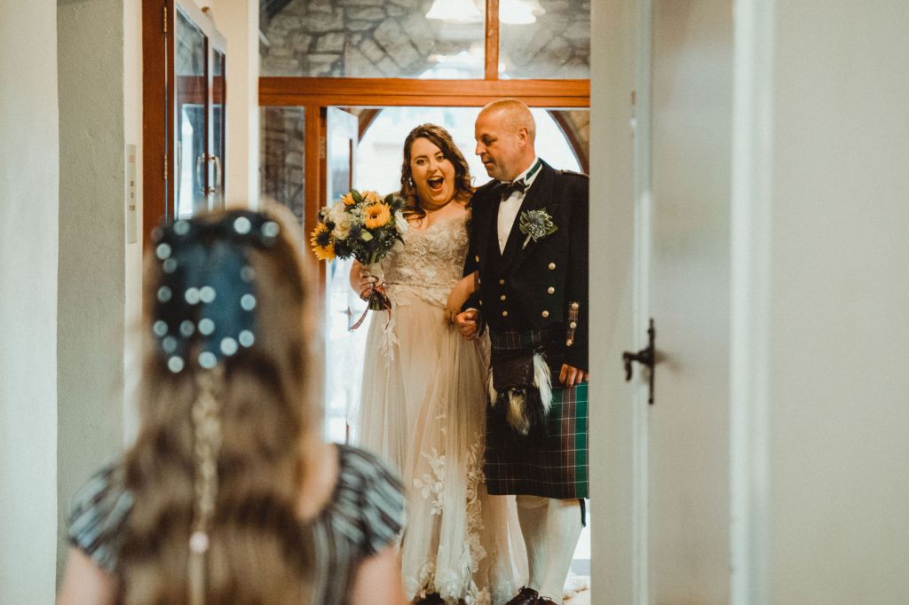 Rosie pulling a funny face at a child, who is out of shot. Rosie is wearing a white wedding dress with lace, and is walking into the church with her dead. 