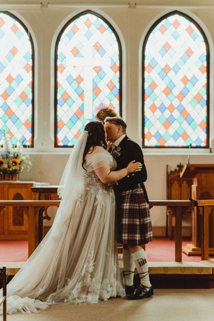 Rosie and Rory kiss in front of three stained glass windows and a wooden alter. She has brown hair and is in a white wedding dress. He has light brown hair and is in a Scottish kilt set.