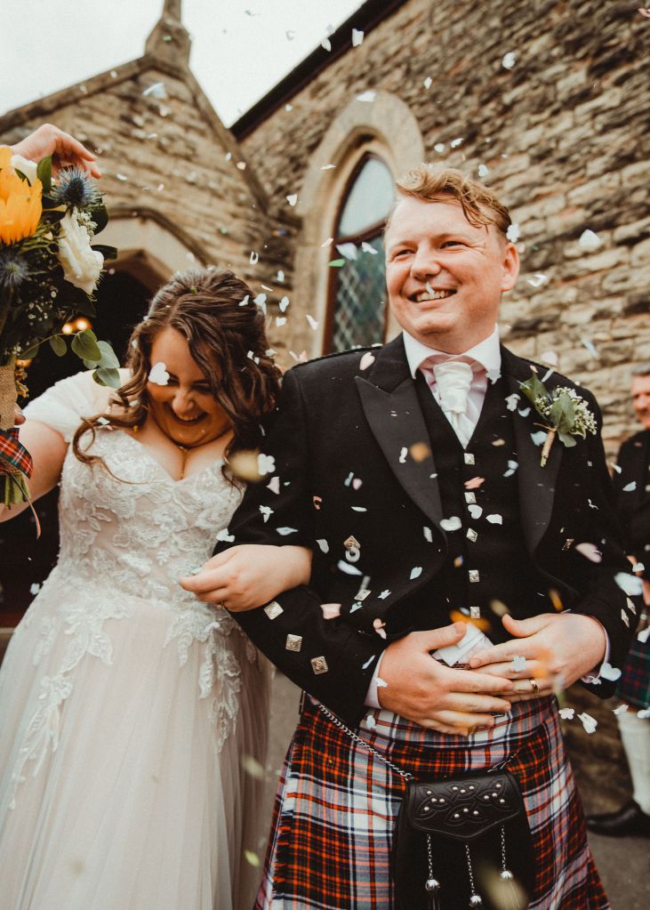 Rosie and Rory leaving the church, arm in arm. There is a brown and grey stone church behind them, and confetti is being thrown all around them.