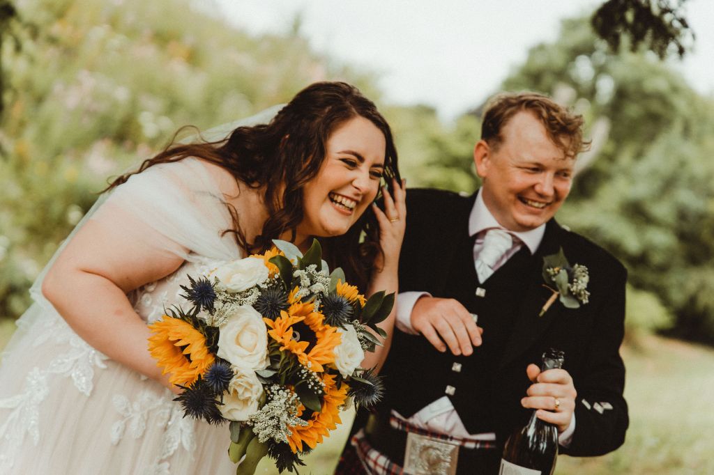 An image of Rosie and Rory grinning at something off camera. On the left, she is holding a bouquet. On the right, he is holding a prosecco bottle. 
