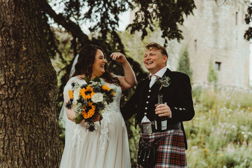 Rosie, on the left on the image, is grinning. She is looking to her left, with her arm up, In her other hand, she carries a bouquet. Rory is to her left, looking off to his right, holding a prosecco bottle in his left hand. 