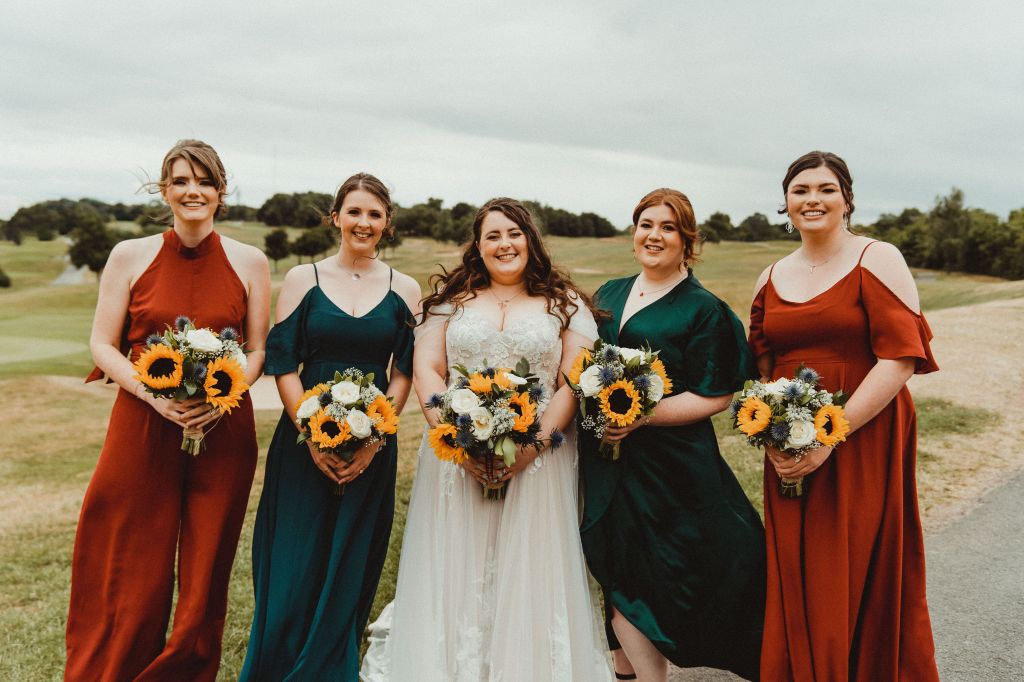 Bridesmaids and bride stood in a row, smiling at the camera, holding bouquet. L-r, red dress, green dress, bride, green dress, red dress.