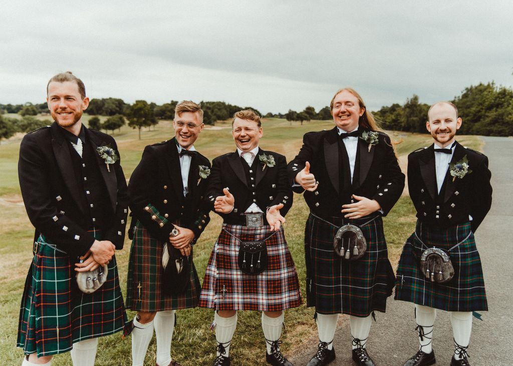 Groomsmen and groom in a row, looking at something to their right and laughing. They are all dressing in traditional kilts. 
