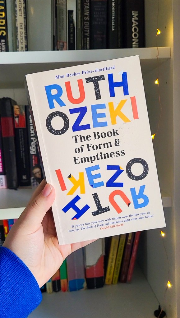 The cover of The Book of Form & Emptiness by Ruth Ozeki, held by Rosie's hand in front of a white bookshelf. The book cover is cream, with the author's name is block capitals in primary colours.