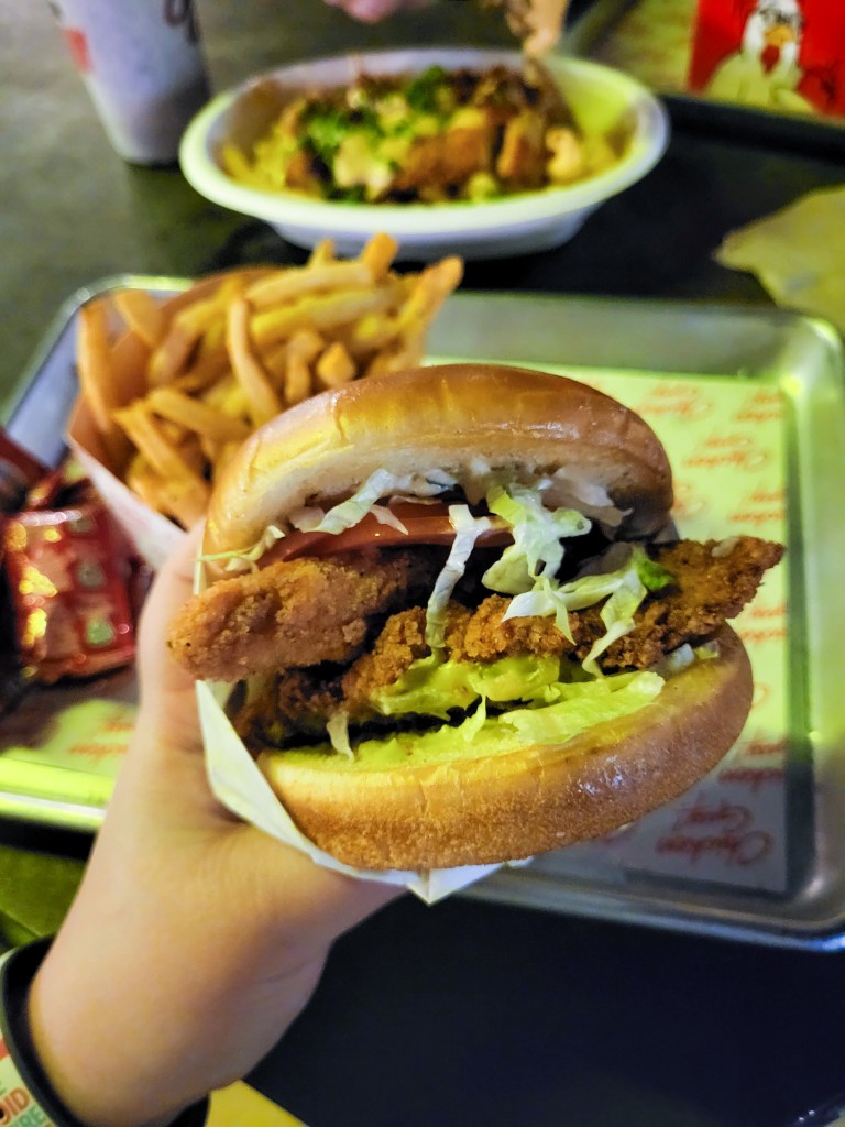 An image of Rosie's hand holding a chicken burger. The burger is made of two buns, two fried chicken pieces, lettuce, tomato, and a sauce. In the background and out of focus, you can see some friends and a mac and cheese basket. 