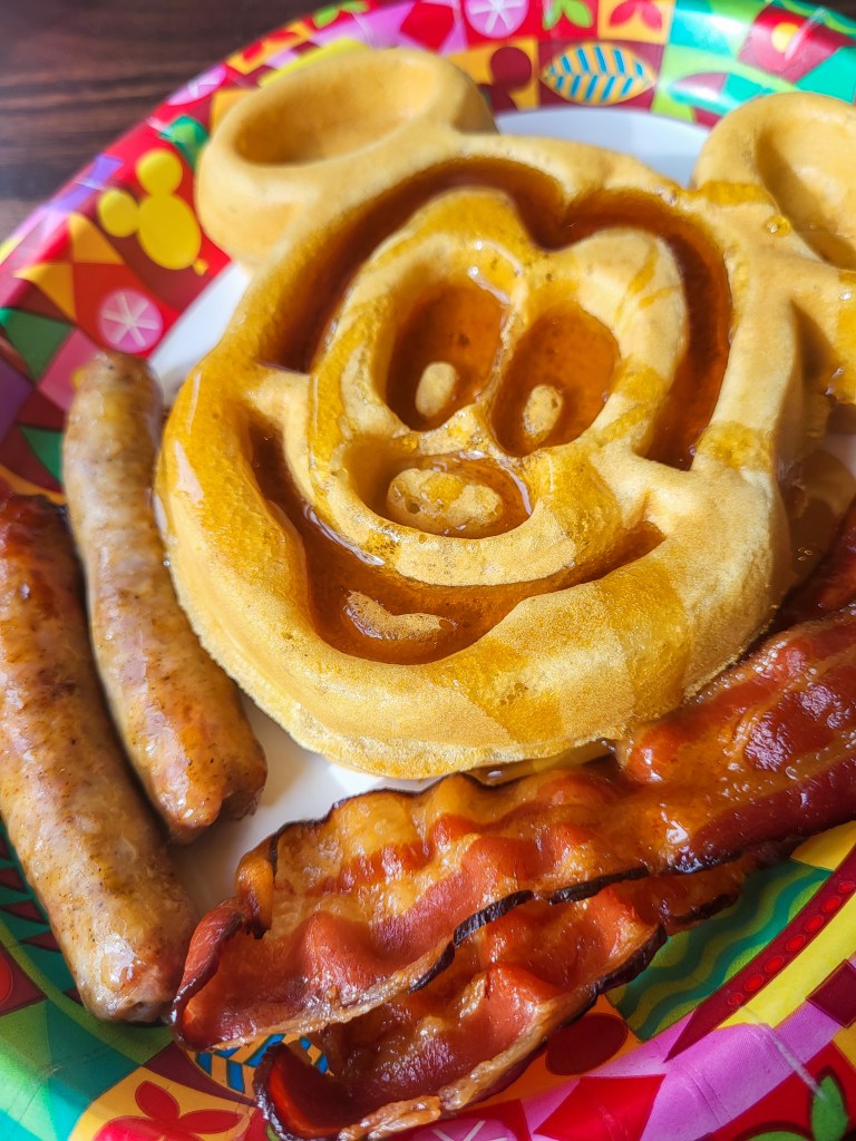 An image of a large Mickey waffle covered in maple syrup, with two sausages and two pieces of bacon. 