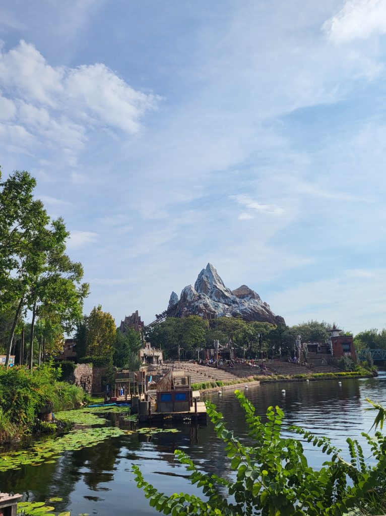 An image of a river leaning to the right, with water lilies and greenery. Behind the river is a man-made snow topped mountain protruding, with a rollercoaster track going into the mountain. The sky is blue with white clouds. 