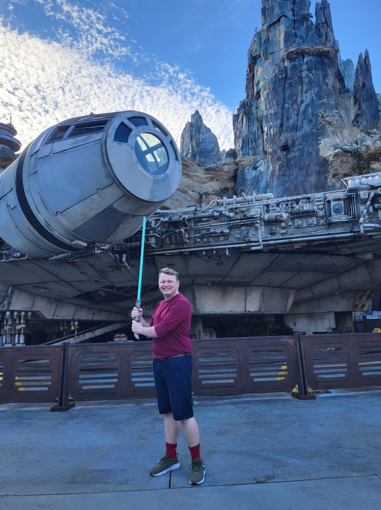 An image of Rory holding a green lightsaber. He is a white man with short, light brown hair, and is wearing a red long sleeved t shirt, blue denim shorts, and green trainers. He is stood in front of a life size Millennium Falcon