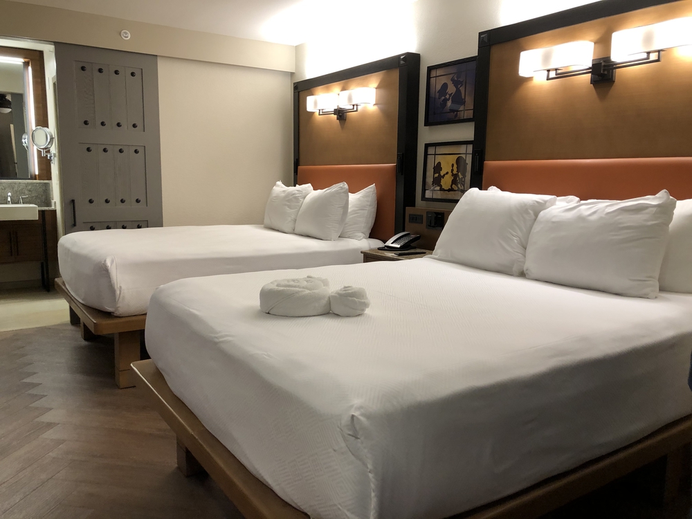 AN image of a standard bedroom at Coronado Springs. The walls are cream, and there are two white, queen sized beds. Each bed has a brown and orange headboard with lights above. There are tonal pictures between the beds and a towel in the shape of Mickey Mouse on the bed. 